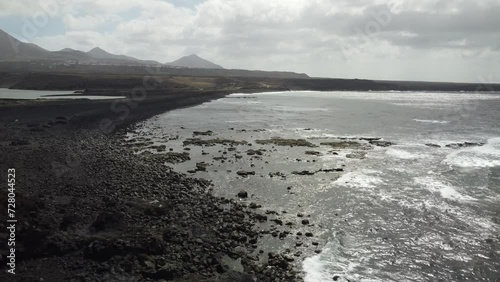 drone shot of a descent from above on black beach with clouds passing and ocean and mountains. 
lanzarote, spain