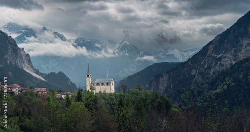 Panoramic view of moody landscape with small church in Dolomites mountains, Belluno, Veneto, Italy. Chiesa Parrocchiale di San Martino in Italian Alps in foggy and cloudy day at springtime