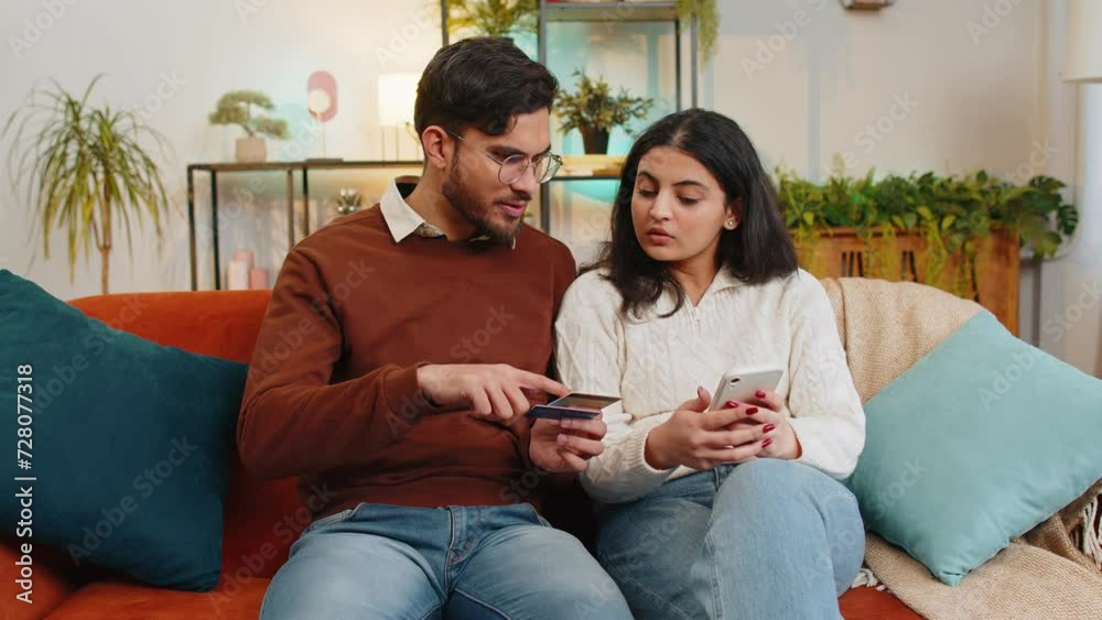 Happy young couple together doing online shopping by entering credit card number on smartphone while sitting on sofa at home. Smiling family are satisfied with a good promotion on goods in apartment. Happy young couple together doing online shopping by entering credit card number on smartphone while sitting on sofa at home. Smiling family are satisfied with a good promotion on goods in apartment.