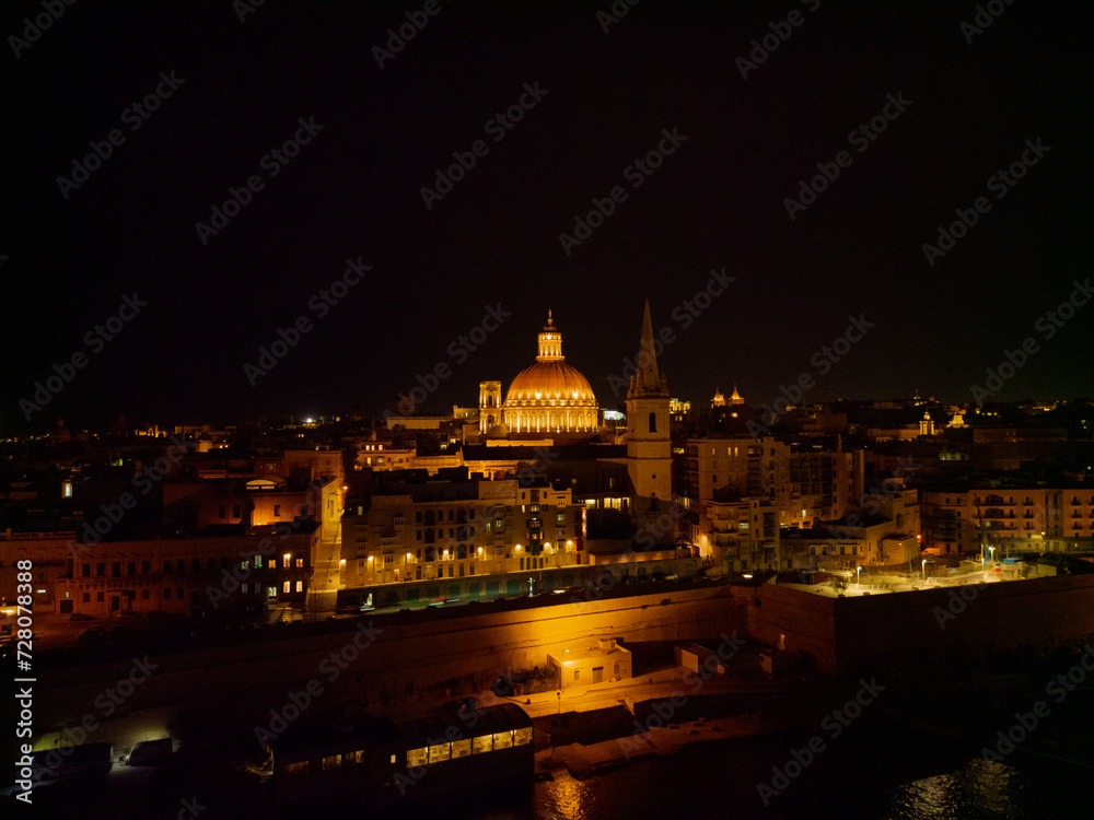 Fototapeta premium Illuminated Basilica dome next to St. Paul's Cathedral at dark night. Aerial view from the sea towards Valletta old town. Residential buildings and streets with street lights.