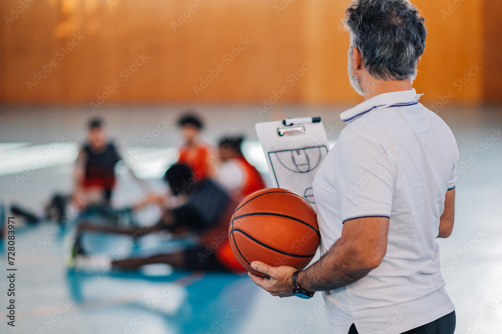 Naklejka premium Rear view of a basketball coach with ball and clipboard in hands on court.