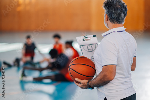 Rear view of a basketball coach with ball and clipboard in hands on court.