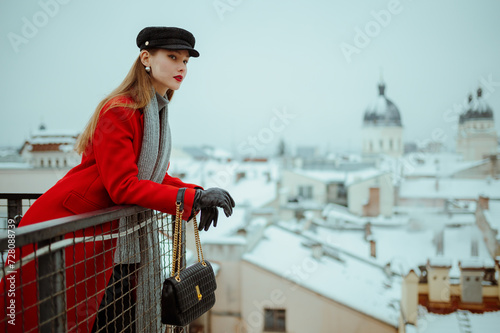 Fashionable woman wearing cap, red winter coat, gray scarf, gloves, holding black leather bag, posing on balcony with beautiful view on snow covered European city. Copy, empty, blank space for text