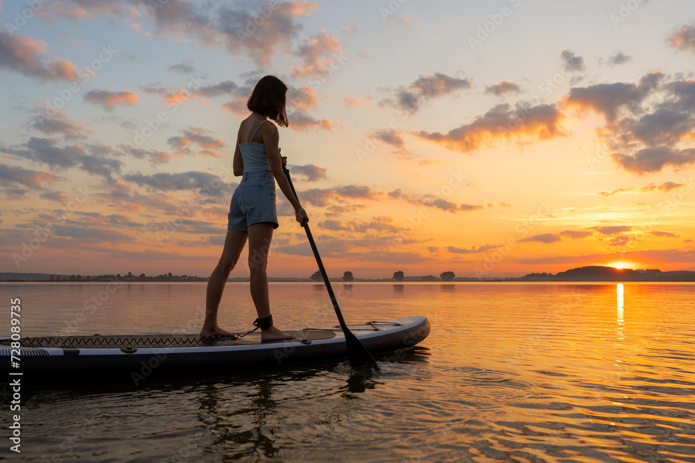 athletic woman on SUP paddle boarding through shining water at sunset. aesthetically wide shot. Freedom happy female relaxing on surfboard on a lake