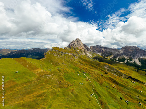 Rocky Seceda ridgeline overgrown with green grass on sunny day. Light clouds and giant forestry mountains around valley in Italian Alps aerial view