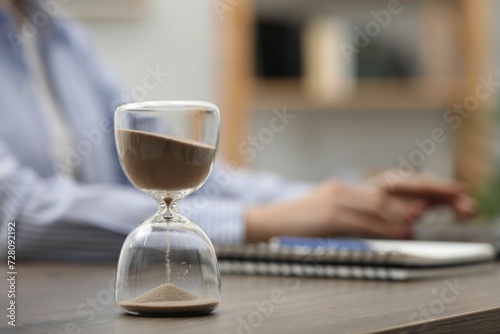 Hourglass with flowing sand on desk. Woman using laptop indoors, selective focus