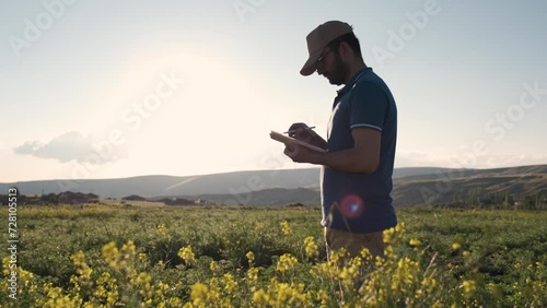 Young farmer, business owner looks in tablet in canola farm land. Young agronomist with tablet in hands. Farmer agronomist checks eco-crops