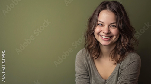 A delighted, successful woman in casual clothing, smiling broadly at the camera, with an olive green background enhancing her confident aura.