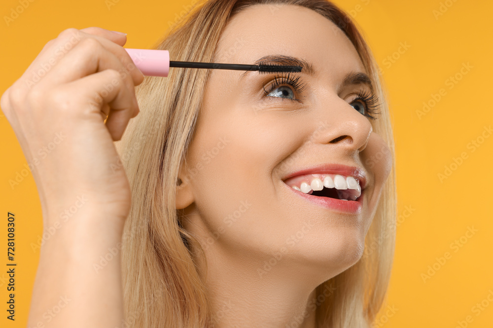 Beautiful woman applying mascara on orange background, closeup