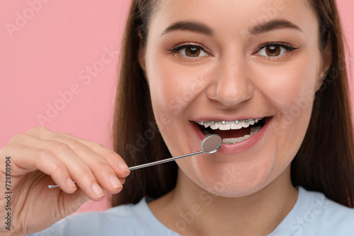 Smiling woman with braces holding dental mirror on pink background  closeup