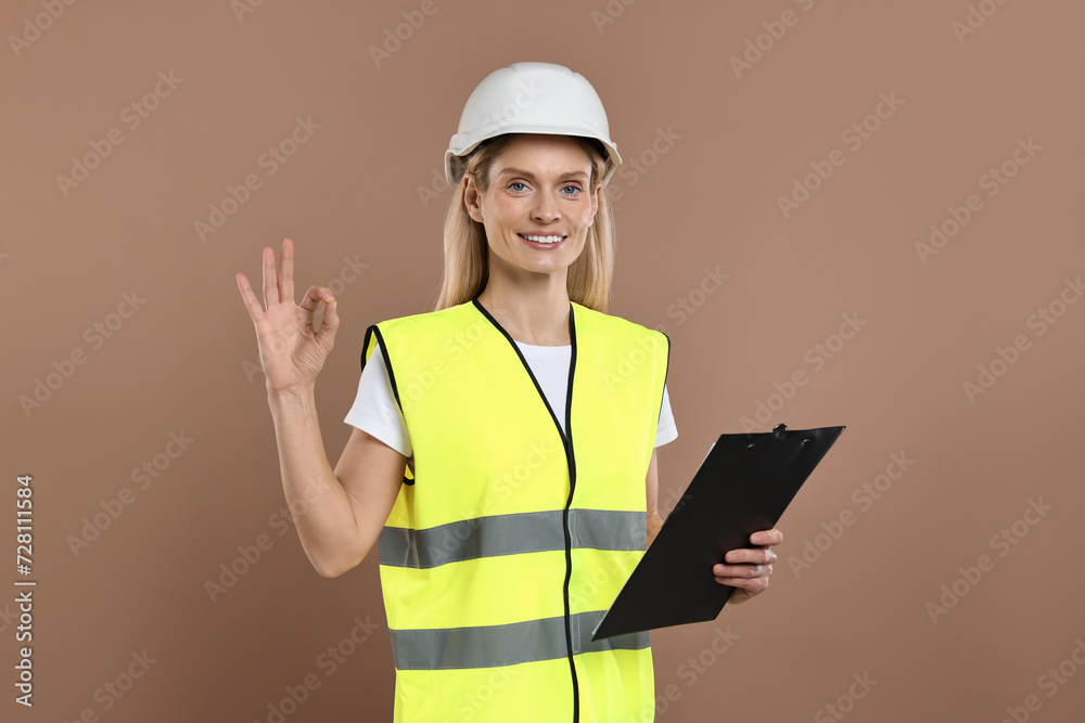 Engineer in hard hat holding clipboard and showing ok gesture on brown background