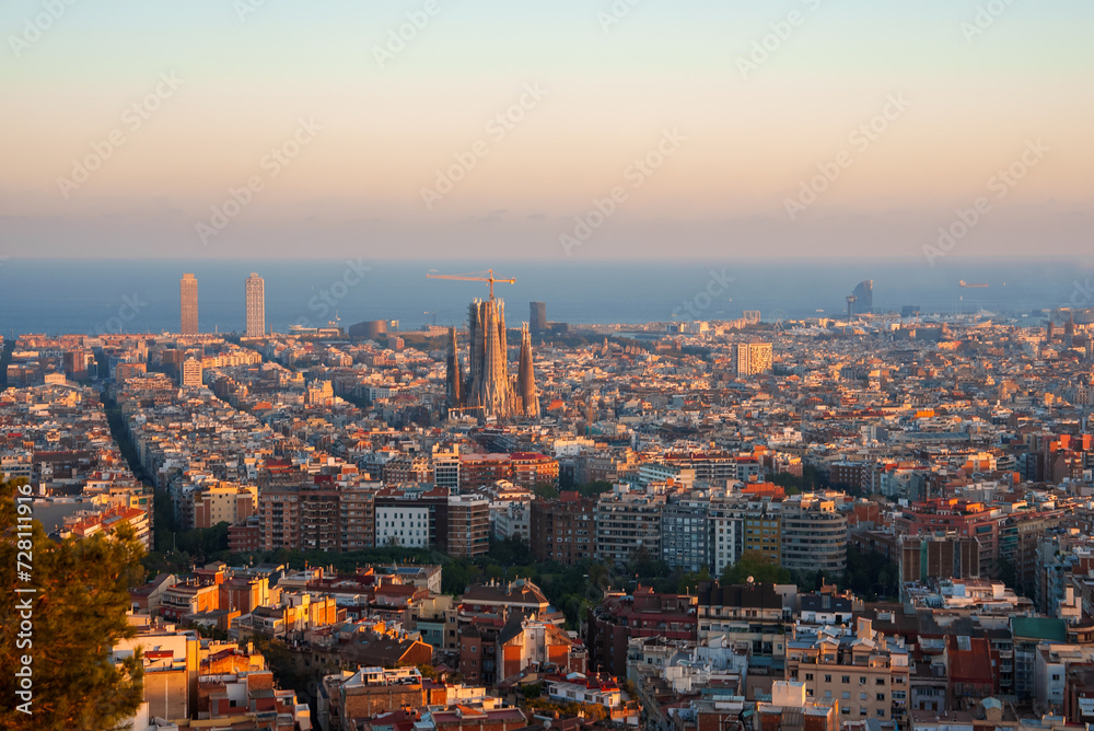 Panoramic view of Barcelona at sunrise or sunset with golden light over the cityscape, featuring the Sagrada Familia and prominent skyscrapers like Torre Glories and Hotel Arts.