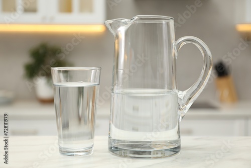 Jug and glass with clear water on white table in kitchen, closeup