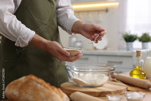 Fototapeta Naklejka Na Ścianę i Meble -  Making bread. Man putting dry yeast into bowl with flour at wooden table in kitchen, closeup