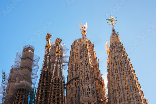 Closeup of Sagrada Familia's spires against a blue sky in Barcelona, showcasing Gaudi's Modernisme style with ornate stonework, sculptures, and a crosstopped tower amid ongoing construction.