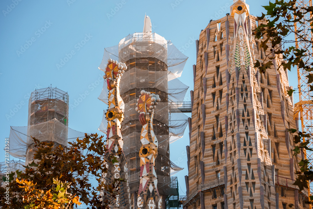 Closeup of Sagrada Familia's towers in Barcelona, adorned with Gaudi's signature floral and geometric patterns, under clear blue skies with visible green foliage.