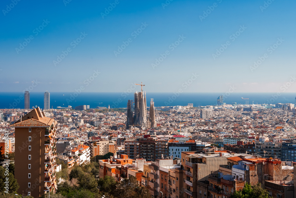 Panoramic view of Barcelona's cityscape featuring the unfinished Sagrada Familia under a clear blue sky, with a backdrop of the Mediterranean Sea and a mix of architectural styles.