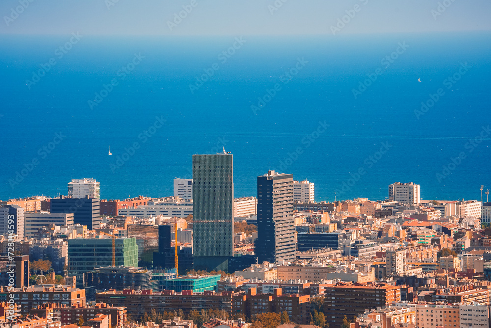 Panoramic view of Barcelona, Spain, highlighting its dense, colorful buildings and modern skyscrapers, with the Mediterranean Sea in the background under clear skies.