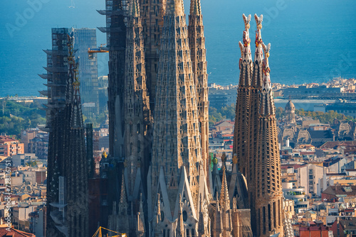 Closeup of Sagrada Familia's spires in Barcelona, showcasing Gaudi's Gothic and Art Nouveau design, with cranes indicating ongoing construction against a cityscape backdrop. photo