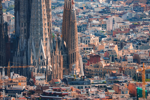 Closeup view of Barcelona's Sagrada Familia, highlighting its intricate spires and ongoing construction against a backdrop of the city's dense, colorful urban landscape in warm afternoon light.