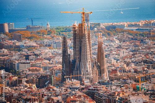 Panoramic view of Barcelona featuring the Sagrada Familia's spires against a cityscape backdrop, with the Mediterranean Sea and clear skies in the distance.