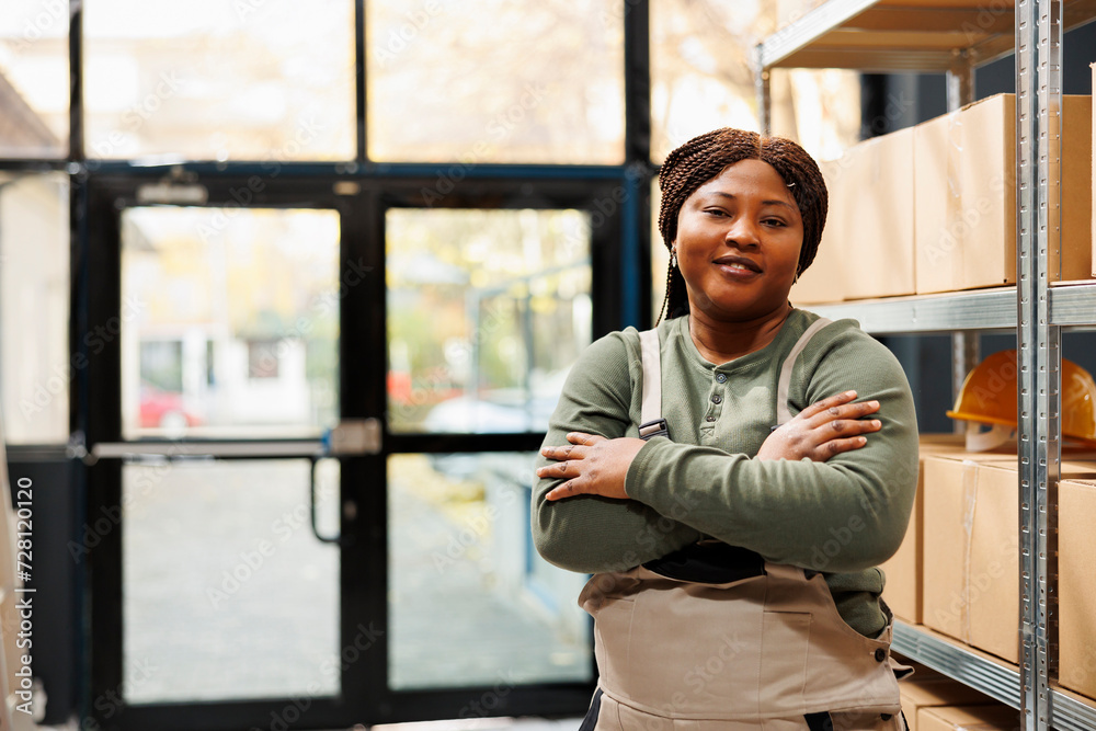 Smiling stockroom manager standing with arm crossed, posing with confidence in warehouse. African american employee wearing industrial overall while working at customers online orders Smiling stockroom manager standing with arm crossed, posing with confidence in warehouse. African american employee wearing industrial overall while working at customers online orders