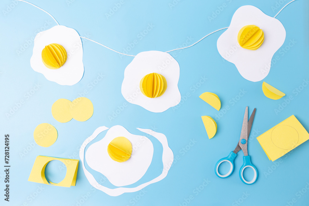 three fried eggs paper craft hanging from a string on a blue background, easter decoration concept, 