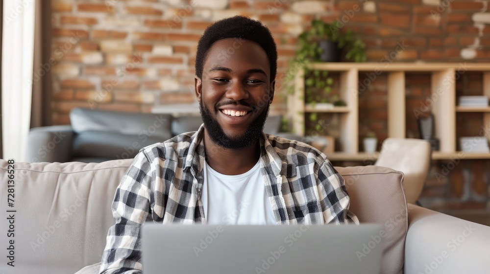 Smiling young black man working or communicating online, sitting on couch with laptop at home. Cheerful millennial guy freelancing, chatting on internet. Modern technologies in everyday life concept Smiling young black man working or communicating online, sitting on couch with laptop at home. Cheerful millennial guy freelancing, chatting on internet. Modern technologies in everyday life concept