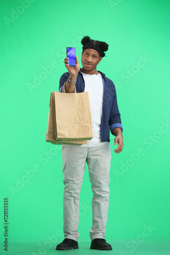 A man, full-length, on a green background, with bags, shows a phone