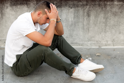 Handsome young man with lot of problems sitting on the ground near the wall holding his head. Depression