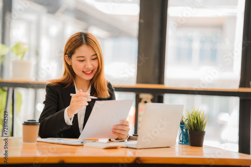 Cheerful Asian woman working with laptop in office, happy in formal suit working in office Charming smiling female office worker, financial accounting concept