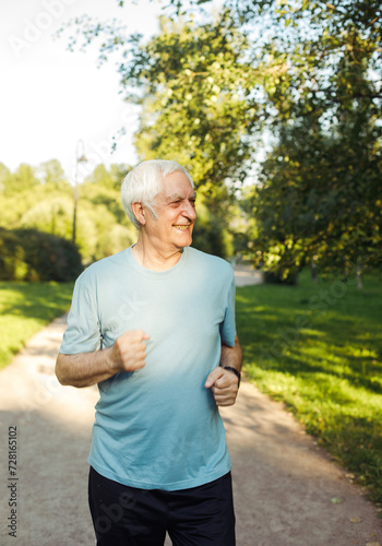 An elderly man in a blue T-shirt is jogging through a summer park.