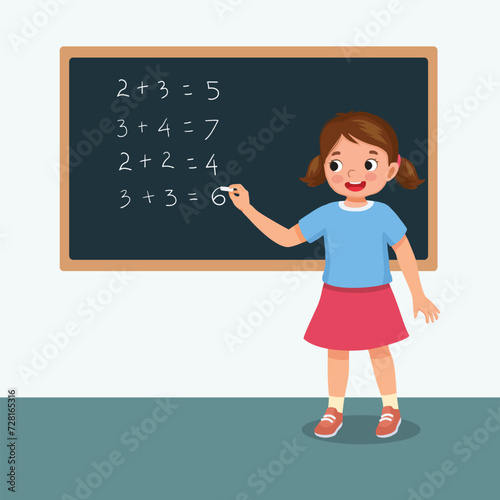 Cute little girl student write on blackboard math equation in front of class with chalk
