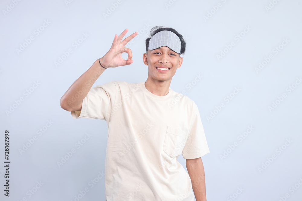 Young man wear pajama sleep and eye mask showing ok sign with finger while looking at camera isolated on white background Young man wear pajama sleep and eye mask showing ok sign with finger while looking at camera isolated on white background