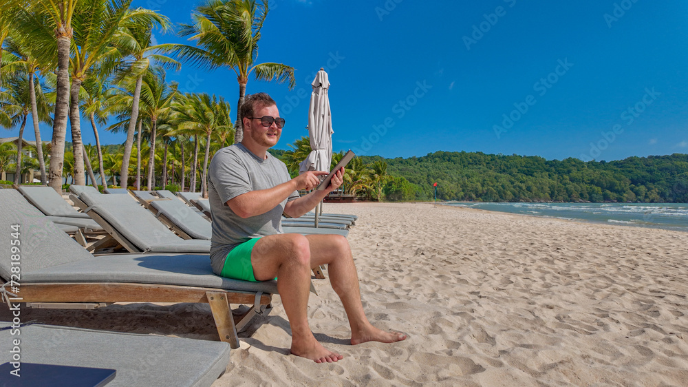 Man using a tablet while relaxing on a sun lounger at a tropical beach resort