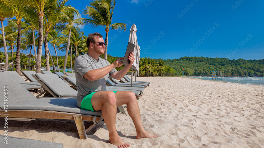 Man in casual summer attire sitting on a beach lounge chair, reading on a tablet, with sandy beach and palm trees in the background