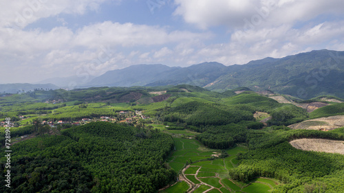 Aerial view of lush green rolling hills and agricultural fields with a small village nestled among the trees, under a cloudy sky background