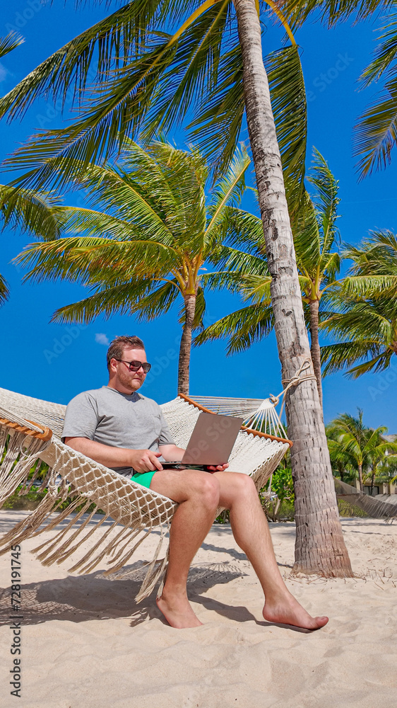 Man working remotely on a laptop while relaxing in a hammock between palm trees on a sunny beach, depicting remote work or digital nomad lifestyle