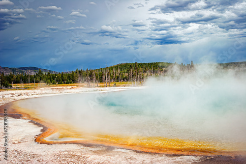 Colorful Yellowstone National Park geyser with hot steam, hillside trees and storm clouds, Wyoming, USA. 