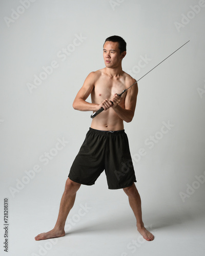 Full length portrait of fit handsome asian male model, wearing gym shorts and shirtless. Holding sword weapons whilst in warrior training action poses, isolated on white studio background.