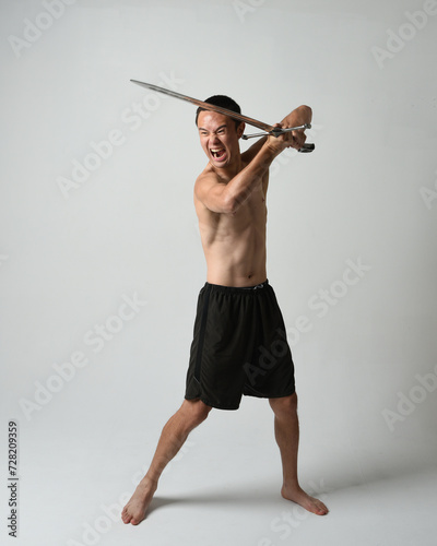 Full length portrait of fit handsome asian male model, wearing gym shorts and shirtless. Holding sword weapons  whilst in warrior training action poses, isolated on white studio background.