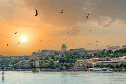 Szechenyi Chain bridge over the Danube River in the city of Budapest. Urban landscape with old buildings, St. Stephens Basilica and opera domes. Reddish sky and flying birds in the background. Hungary