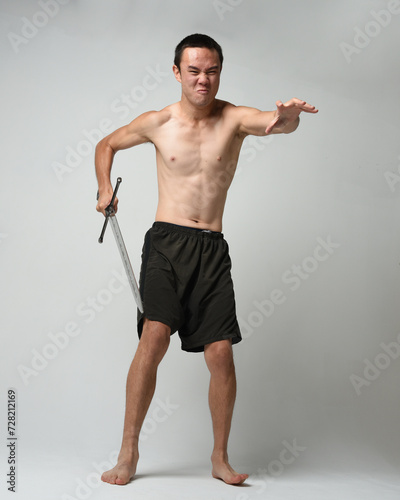 Full length portrait of fit handsome asian male model, wearing gym shorts and shirtless. Holding sword weapons whilst in warrior training action poses, isolated on white studio background.
