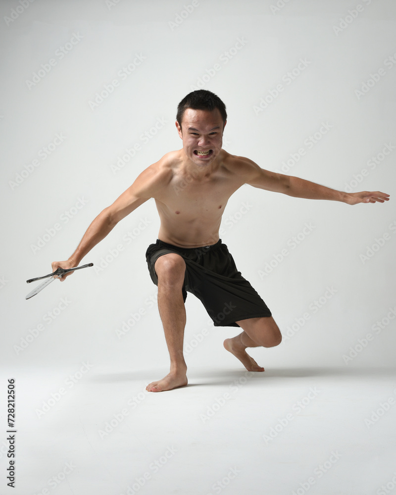 Obraz premium Full length portrait of fit handsome asian male model, wearing gym shorts and shirtless. Holding sword weapons whilst in warrior training action poses, isolated on white studio background.