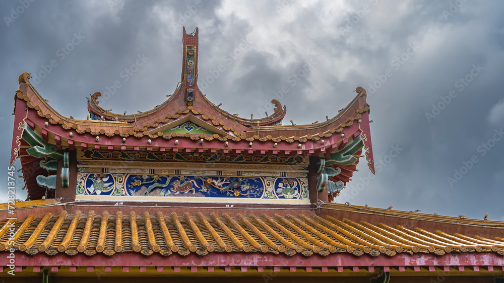Details of the architecture of the Chinese Thean Hou Temple. The tiled roof with curved edges is decorated with carved ornaments and drawings. The background is a cloudy sky. Malaysia. Kuala Lumpur.