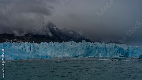 The amazing Perito Moreno Glacier. A wall of blue ice with cracks and sharp peaks stretches in a glacial lake. Iceberg and melted ice floes float in turquoise water. Mountains in clouds and fog. 