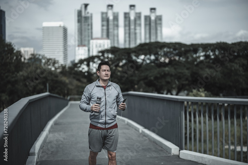 A man holding a water bottle, smartphone and listen music earphone while running up on footbridge in the city center park for cardio workout. Health and Lifestyle in big city life concept.