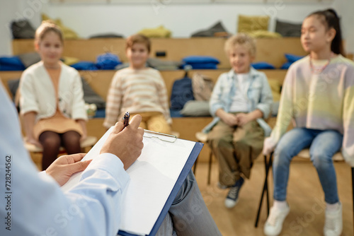 Close up of teacher or therapist holding clipboard and taking notes while working with group of childrenwith copy space