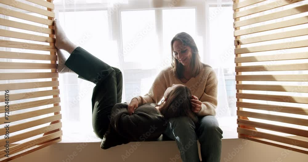 Two lesbian woman enjoy spend time together, sitting in embrace and talking at home. Female couple in romantic relationship. LGBT partners relaxing together on window sill Two lesbian woman enjoy spend time together, sitting in embrace and talking at home. Female couple in romantic relationship. LGBT partners relaxing together on window sill