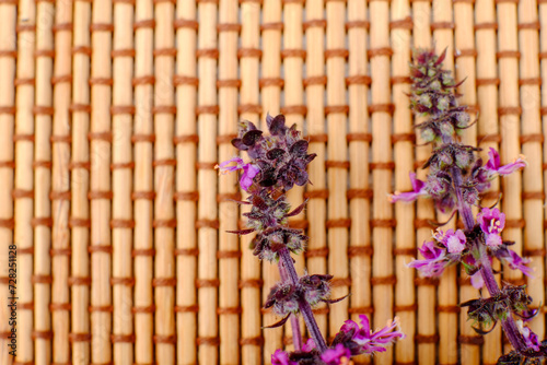 This image presents two sprigs of lavender with delicate purple flowers and a deep greenish-purple hue to the leaves, lying atop a traditional woven mat. The mat's texture contrasts with the organic